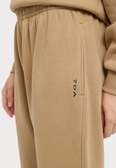 Beige sweats met een elastische tailleband, zijzakken en een marineblauw geborduurd "7DA" logo nabij de heup. Zachte, gestructureerde stof.