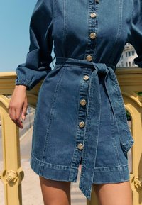 Denimkleit, millel on tihedalt istuv ülaosa, pikad volangvarrukad ja vööümber seotav pael. Kaunistatud kuldse tooniga nööpidega ning veidi lehviva seelikuga.