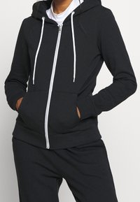 Felpa nera con zip realizzata in tessuto morbido, con zip bianca, cordini e tasche frontali. Vestibilità slim con polsini e orlo a coste.