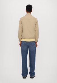 Giacca di cotone beige con colletto, inserto posteriore e polsini con bottoni, abbinata a jeans blu larghi e scarpe nere.