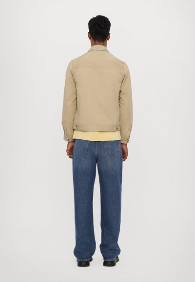 Beige cotton jacket with a collar, back yoke, and buttoned cuffs, paired with blue loose-fitting jeans and black shoes.
