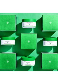 Grüne Kästen und runde Gläser mit grünen Deckeln, die auf einer leuchtend grünen Oberfläche angeordnet sind. Etiketten zeigen Produktnamen in weißem Text.