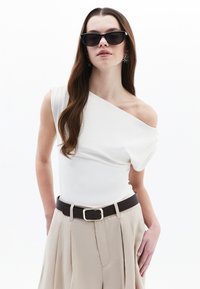 Top bianco scollato con dettagli drappeggiati, abbinato a pantaloni beige a vita alta e una cintura marrone scuro con una fibbia argentata. Occhiali da sole neri.