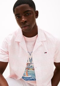 Camicia a maniche corte di colore rosa chiaro con un motivo sottile, indossata sopra una t-shirt grafica di un rosa pastello con un design di una barca. Presente un piccolo dettaglio del logo.