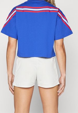 Personne portant un haut court bleu avec des rayures rouges et blanches sur les épaules et un short de sport blanc, vue de dos.