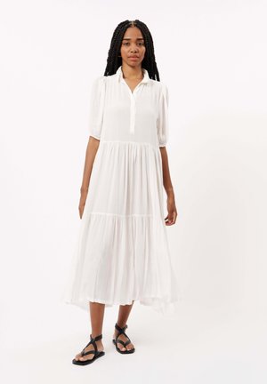 Robe maxi blanche à volants avec de courtes manches bouffantes, col à boutons et une silhouette fluide, associée à des sandales plates noires.