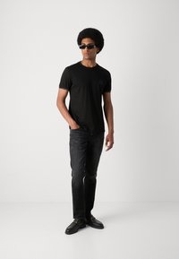 Zwart t-shirt met korte mouwen en een borstzak, gecombineerd met donkere jeans en zwarte instappers. Accessoires zijn onder andere donkere zonnebrillen.