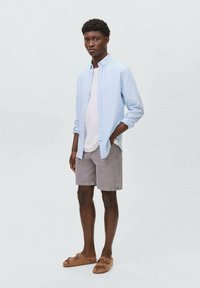Un giovane uomo si trova contro uno sfondo grigio chiaro, indossando una camicia azzurra con bottoni, una maglietta bianca, pantaloni corti grigi e sandali marroni.