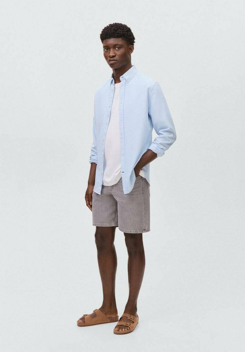 Un giovane uomo si trova contro uno sfondo grigio chiaro, indossando una camicia azzurra con bottoni, una maglietta bianca, pantaloni corti grigi e sandali marroni.