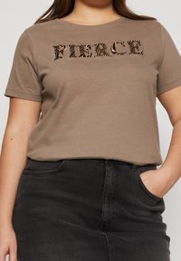 Brun kortärmad t-shirt med texten "FIERCE" i ett strukturerat djurmönster. Buren med en svart jeanskjol.