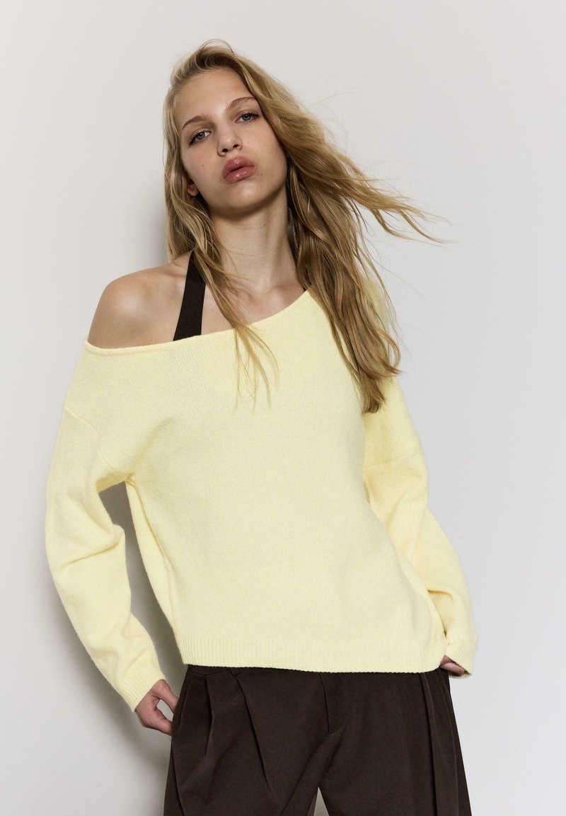 Femme blonde portant un pull ample jaune à épaules dénudées par-dessus un haut dos nu noir, associé à un pantalon large marron foncé, sur un fond uni.