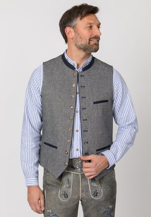 Mann in traditioneller bayerischer Tracht mit einer grauen Wollweste über einem blau gestreiften Hemd und bestickten Lederhosen.