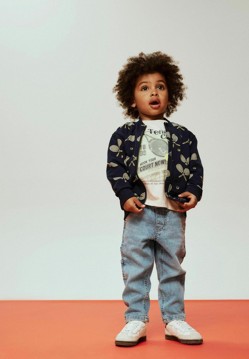 Tamsiai mėlyna bombonešio striukė su geltonu teniso raketės raštu, balta grafine marškinėliai, šviesiai mėlyni platūs džinsai ir balti sportbačiai, stovintys ant raudonos grindys.