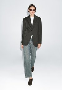 Blazer grigio scuro con vestibilità aderente, indossato sopra un top bianco e jeans a gamba larga azzurri. Mocassini marroni completano il look. Le montature sono scure.