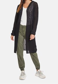 Černý dlouhý kardigan vyrobený z lehkého, průhledného materiálu, s otevřeným předním dílem a dlouhými rukávy, spárovaný s olivově zelenými tepláky a teniskami.