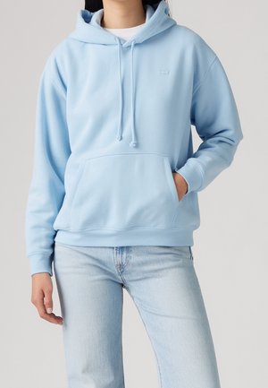 Person wearing light blue hoodie with front pocket and drawstrings, paired with light blue jeans against a plain background.