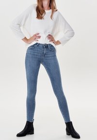 Donna con una maglietta bianca a maniche lunghe, jeans skinny blu a vita alta e stivaletti neri, in piedi con le mani sui fianchi.