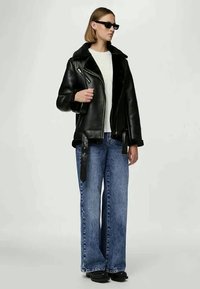 Veste de moto en cuir noir avec un col en shearling, associée à un jean large bleu clair et des chaussures noires. Lunettes de soleil comprises.