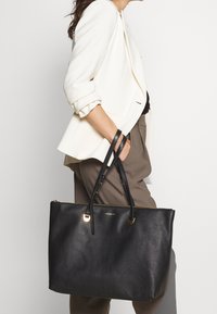 Zwarte leren tote tas met een gestructureerde afwerking, goudkleurige hardware accenten, dubbele handvatten en een ritssluiting. Minimalistisch ontwerp met een ruime binnenkant.