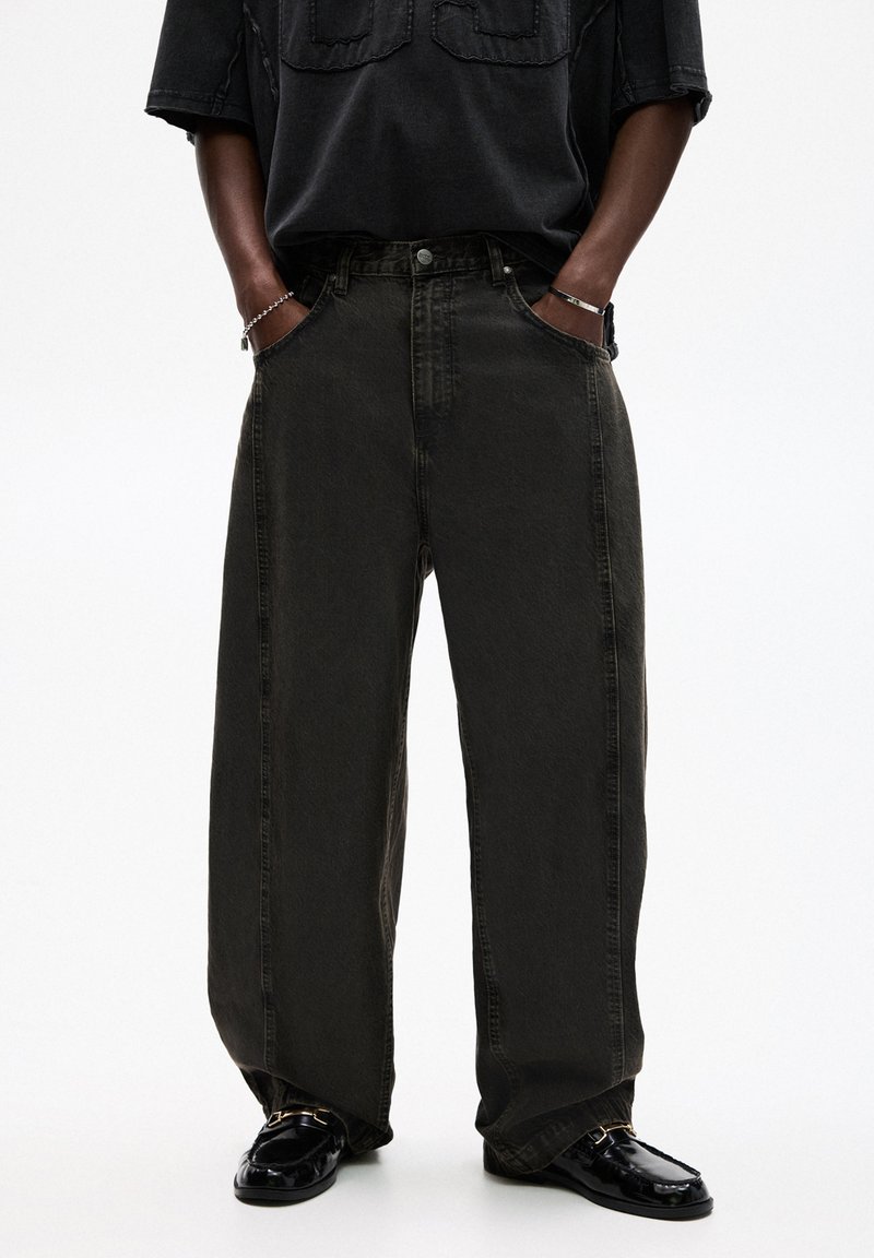 Jeans in denim verde scuro, con vestibilità rilassata e vita alta. Presentano tasche frontali e chiusura con bottone. Indossati con una camicia nera larga e scarpe lucide.
