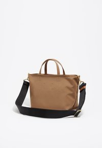 Bolso de tela de color beige con dos asas superiores y una correa negra desmontable. Presenta herrajes dorados y un diseño minimalista. Textura suave.