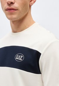 Κοντινό πλάνο ενός ατόμου που φοράει άσπρο πουκάμισο με οριζόντια σκούρα μπλε ρίγα και λογότυπο EA7 Emporio Armani στο στήθος.