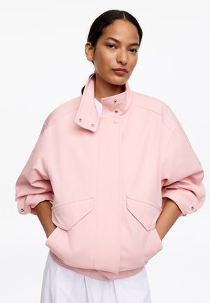 Femme portant une veste ample rose clair avec col montant et manches retroussées, debout les mains dans les poches sur fond blanc.