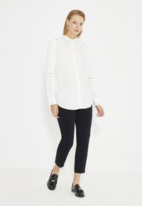Witte blouse met lange mouwen, knoopsluiting en een bandkraag met fijne details. Gepaarde met zwarte tapered broeken en glanzende zwarte instappers.