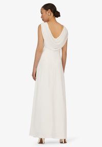 Eine Frau mit einem Dutt, die ein langes, ärmelloses weißes Kleid mit einem drapierten, offenen Rücken trägt, steht und ist mit dem Rücken zu uns auf einem weißen Hintergrund.