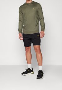 Sudadera verde oliva con puños acanalados, combinada con pantalones cortos negros con un logo amarillo. Se lleva con zapatillas deportivas negras y calcetines blancos.