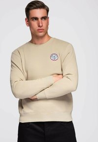 Beige sweatshirt met een ronde hals, geribbelde manchetten en een logopatch op de linkerborst. Zachte stof met een gladde textuur.