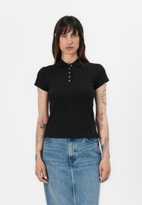 Zwarte poloshirt met korte mouwen en een kraag, afgewerkt met goudkleurige knopen. Gecombineerd met een lichtblauwe spijkerrok.