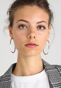 Sidabrinės metalinės žiedinės auskarai su poliruotu paviršiumi, derinami su langeliais raštuotu švarku ir baltu apvalaus kaklo marškinėliais. Švarus, modernus dizainas.