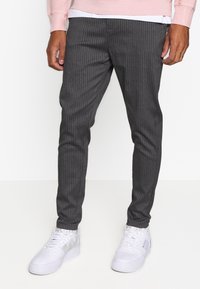 Person wearing slim-fit dark gray pinstripe trousers, white sneakers, and a light pink long-sleeve top, standing against a plain white background.