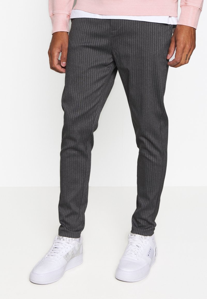 Person wearing slim-fit dark gray pinstripe trousers, white sneakers, and a light pink long-sleeve top, standing against a plain white background.