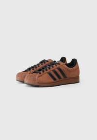 Brūnas zamšādas Adidas Superstar kedas ar melnām svītrām un auklām, novietotas blakus vienai otrai uz vienkārša baltā fona.