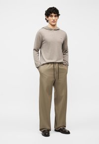 Jeune homme aux cheveux bouclés portant un sweat à capuche beige, un pantalon ample kaki avec cordon de serrage et des chaussures habillées noires, debout les mains dans les poches.