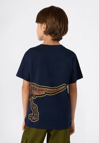 Dziecko z brązowymi włosami nosi granatową koszulkę z pomarańczowo-żółtym wzorem szkieletu dinozaura na plecach, w połączeniu z oliwkowymi spodniami.