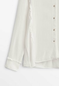 Blouse blanche texturée à manches longues avec bords volantés et petits boutons ronds sur le devant, montrée du milieu de la manche à l'ourlet.