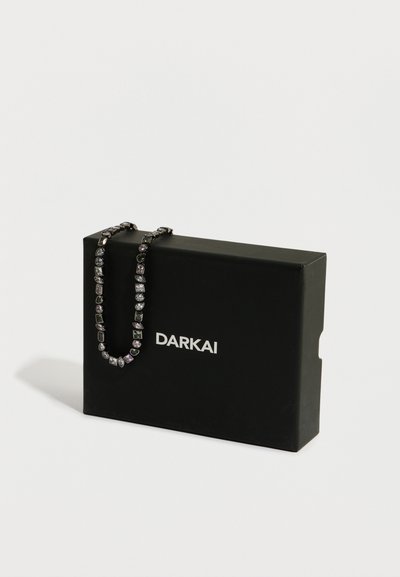 Sort smykkeskrin med "DARKAI"-logo; indeholder en sølvfarvet halskæde prydet med sten i forskellige former i lilla og klare nuancer.