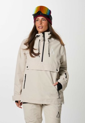 Femme portant une veste et un pantalon de ski beige, un bonnet bordeaux et des lunettes de ski réfléchissantes sur la tête, posant devant un fond uni.