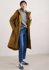 Manteau long vert olive avec capuche, pull gris, jeans bleus et baskets bleues avec rayures blanches. Présente un tissu texturé et une fermeture éclair.