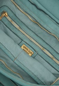 Interno di una borsa in pelle teal con molte cerniere dorate, foderata in tessuto e un'etichetta che recita "Caterina Lucchi, Made in Italy."