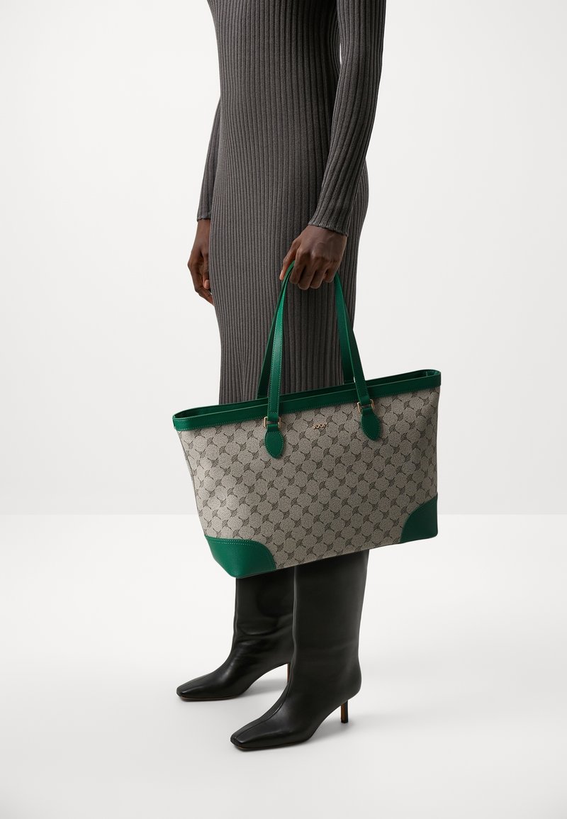 Personne portant une robe grise côtelée tenant un grand sac cabas beige à motif avec des garnitures en cuir vert, portant des bottines noires à talons.