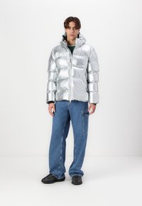 Giubbotto puffer argento con design lucido e imbottito, cappuccio e chiusura con zip. Abbinato a pantaloni in denim blu e sneaker nere. Sfondo semplice.