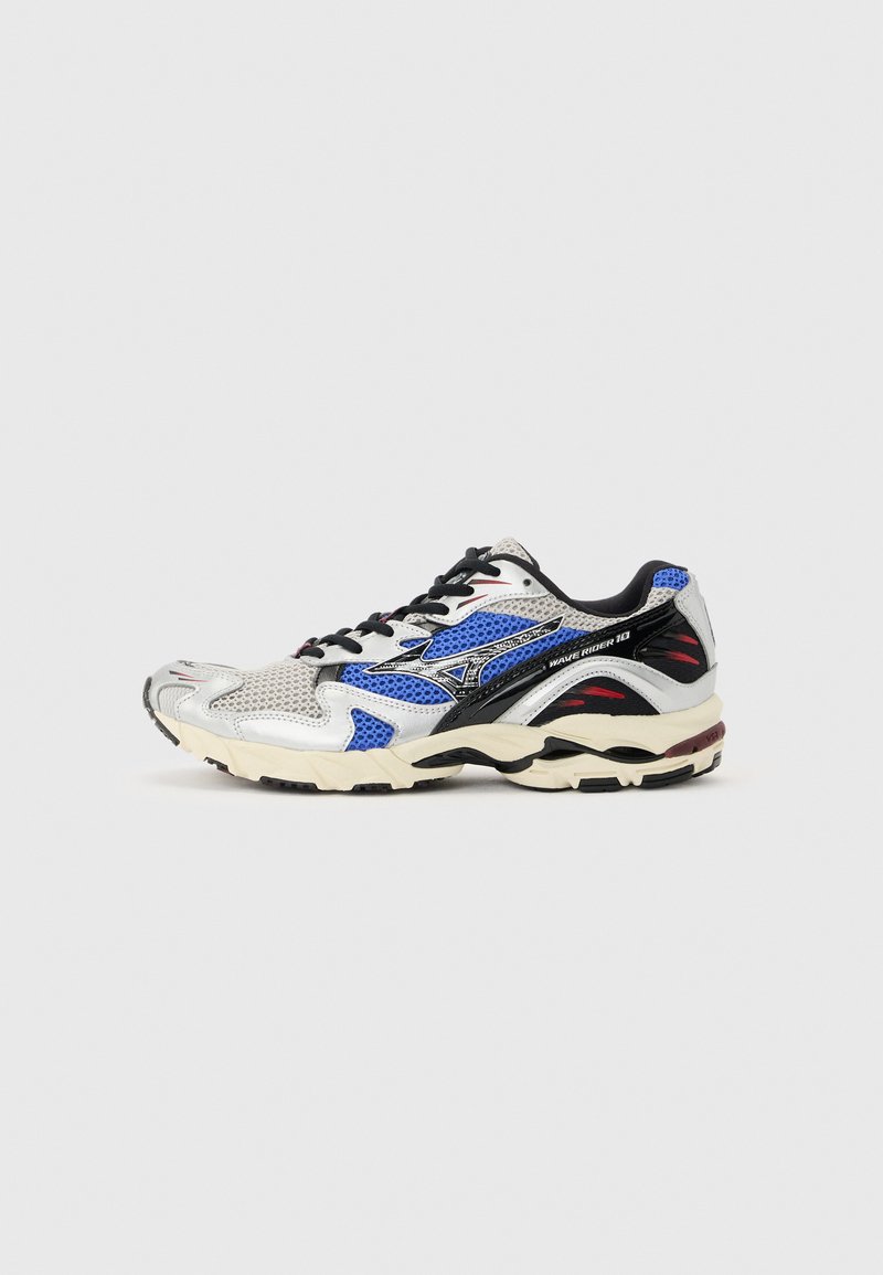 Αθλητικό παπούτσι με πλέγμα και συνθετικά πάνελ σε λευκό, μπλε, μαύρο και κόκκινο, μαύρα κορδόνια και μαλακή μπεζ σόλα σε ανοιχτό γκρι φόντο.