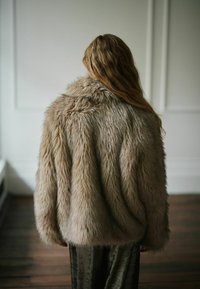 Osoba s dlhými vlnitými vlasmi, ktorá má na sebe nadýchaný béžový kabát z umelej kožušiny, stojí vo vnútri na drevenej podlahe a je otočená chrbtom k fotoaparátu.