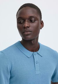 Polo bleu clair en maille avec une texture douce, manches courtes et col classique. Comprend deux boutons au niveau du col.