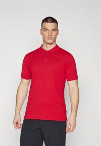 Crvena polo majica od pamuka, s ovratnikom, tri gumba, kratkim rukavima i malim crnim logotipom na prsima.