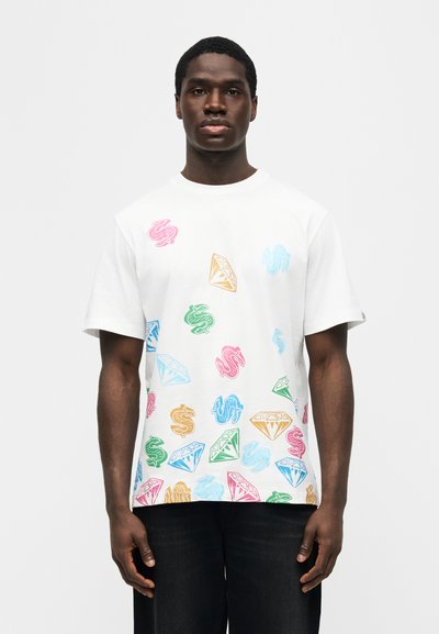 Νεαρός άνδρας που φοράει λευκό κοντομάνικο πουκάμισο με πολύχρωμα σύμβολα δολαρίου και διαμαντιών, συνδυασμένο με μαύρο παντελόνι, στέκεται μπροστά σε απλό φόντο.